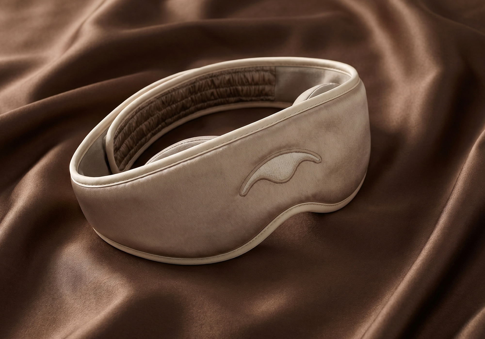 A sepia-toned photo of a cushioned sleep mask resting on a silky brown fabric.