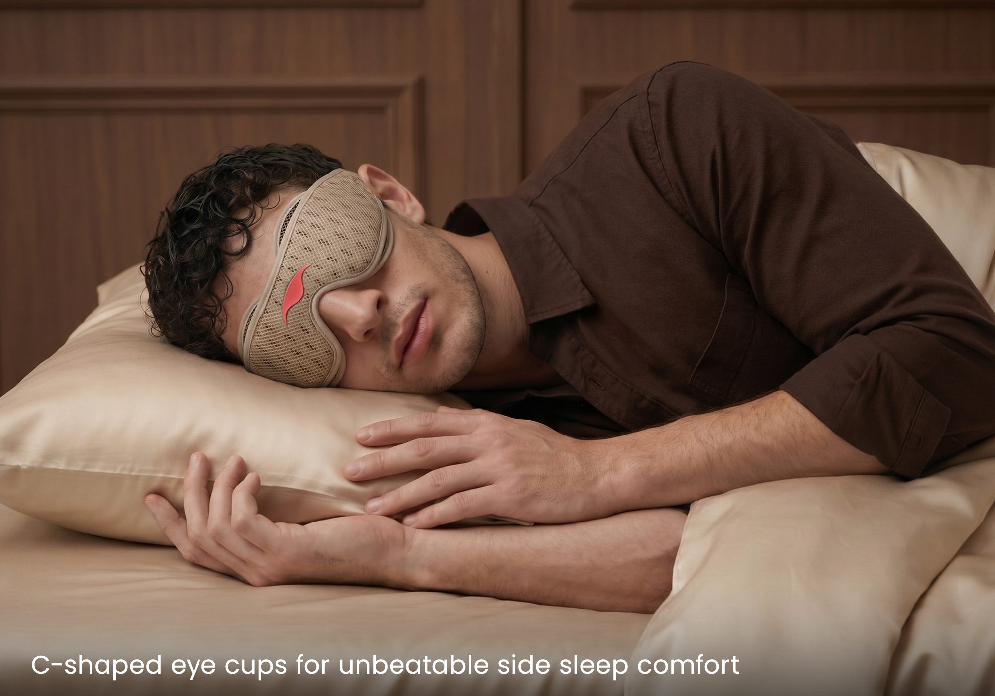 Man wearing a brown sleeping mask with red logo lying down on cream-colored silk pillow.