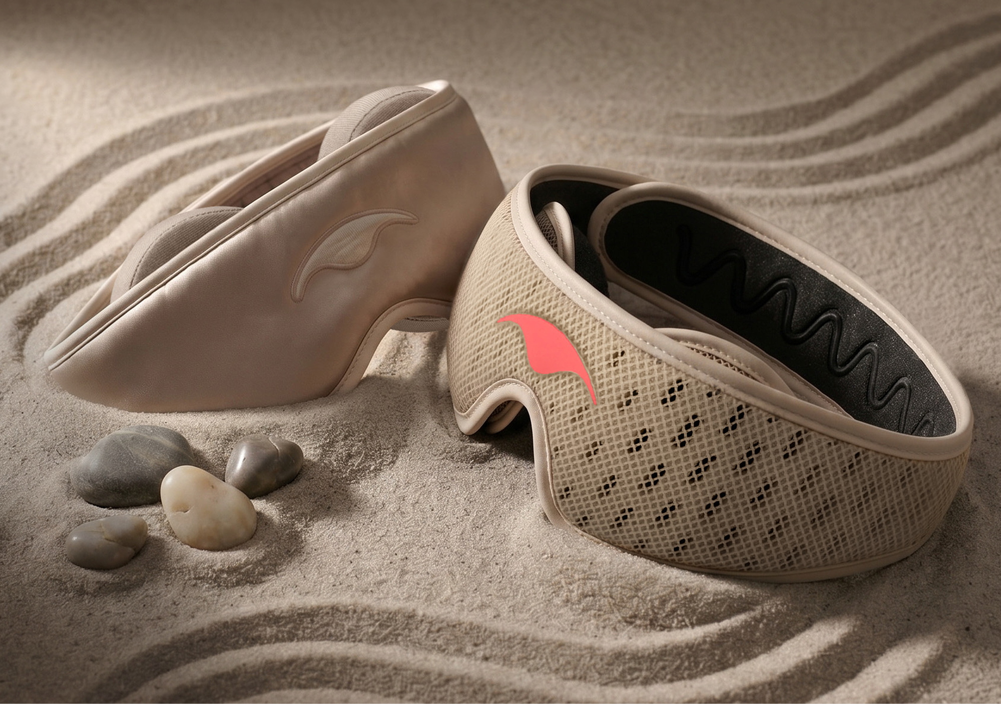 Two beige eye massagers with straps rest on raked sand next to several smooth stones.