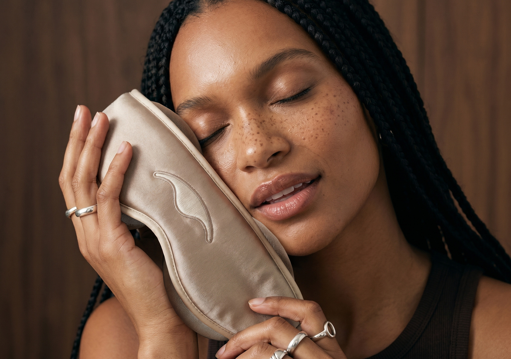 A woman with braided hair and closed eyes holds a light-colored, satin sleep mask against her cheek.