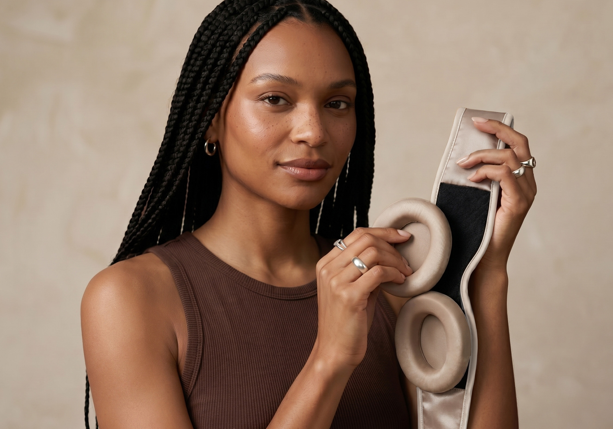 A woman with long braids holds a silk sleep mask with padded eye cups while looking directly at the camera.