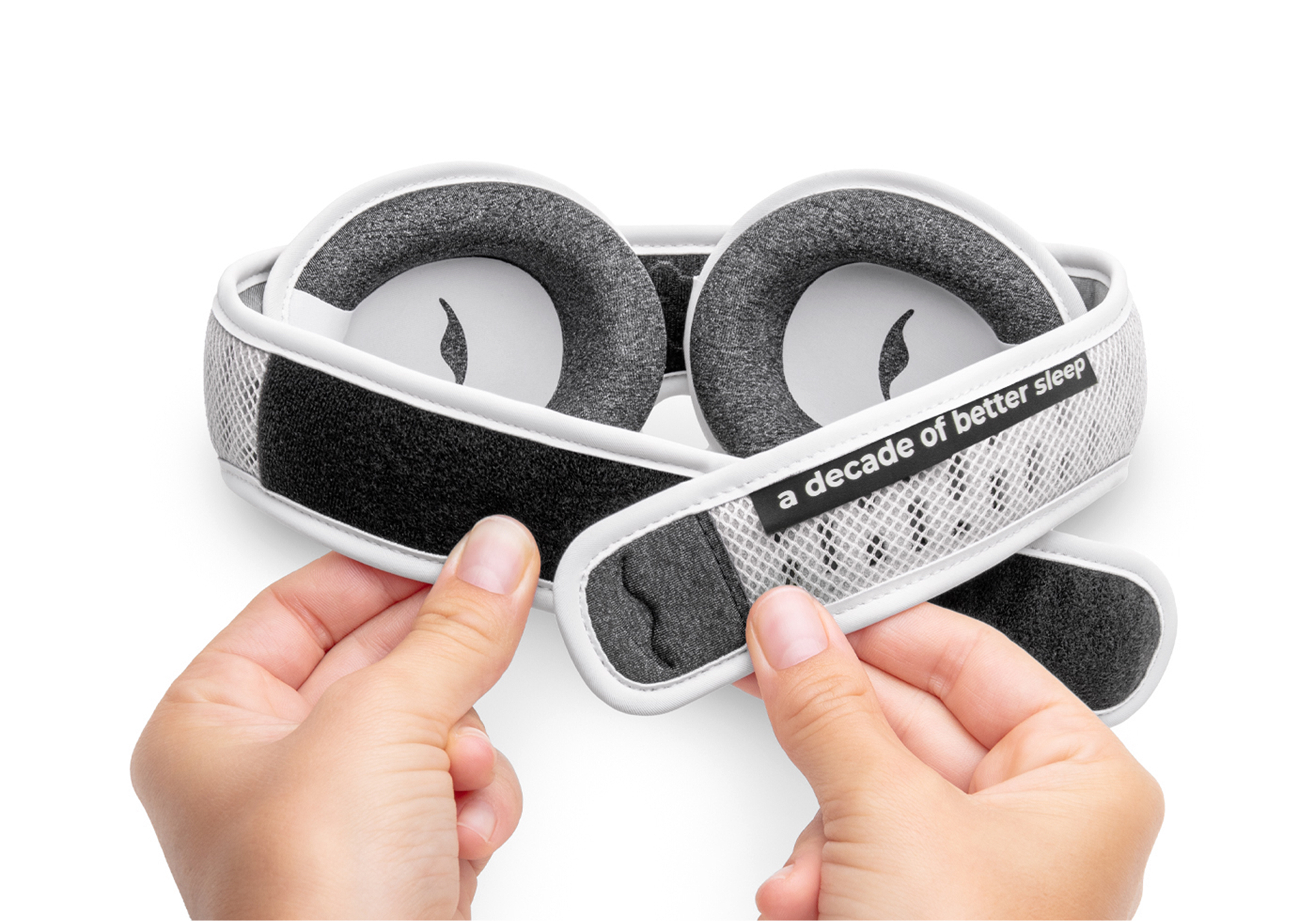 A pair of hands adjusts the velcro strap of a white and grey padded sleep mask.