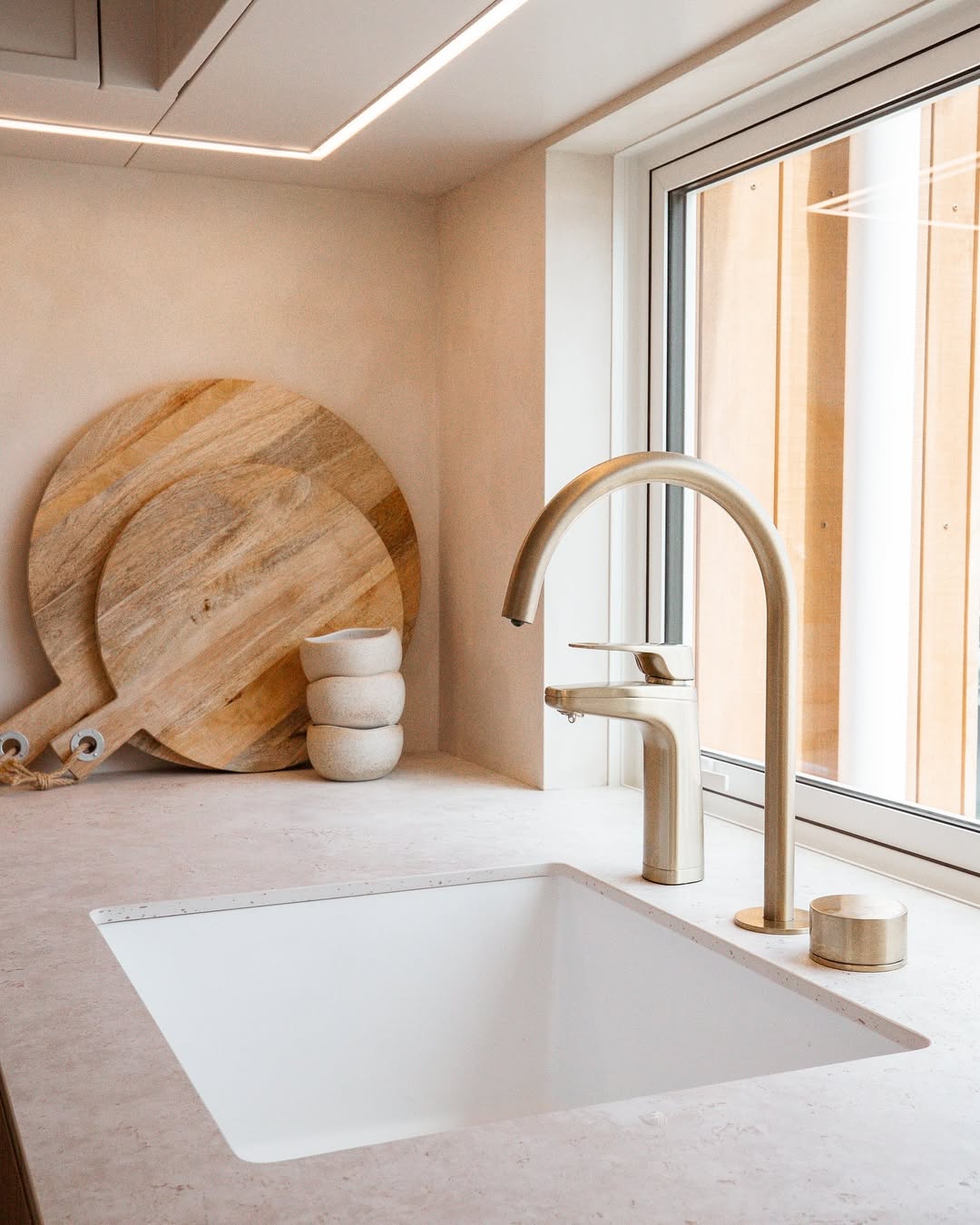Modern kitchen sink with brass faucet, cutting boards, and stacked bowls by a window.