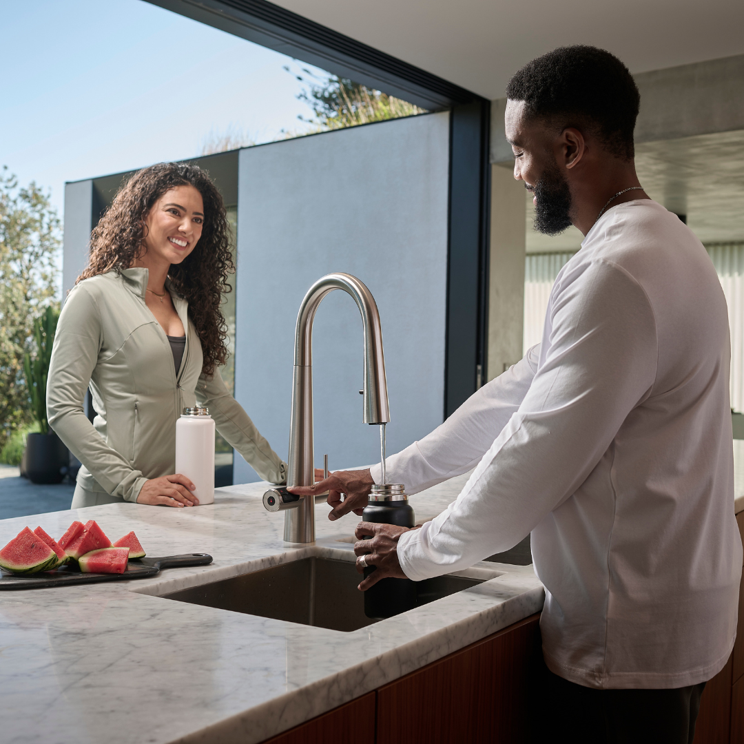 Two people in a kitchen, one filling a bottle at the sink, with watermelon on the counter.