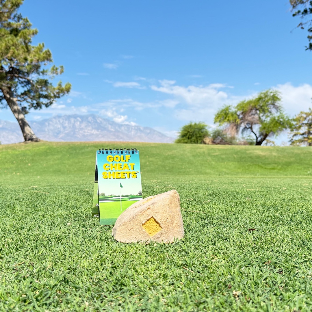 A 'Golf Cheat Sheets' booklet and a yellow tee marker on a sunny golf course with mountains.