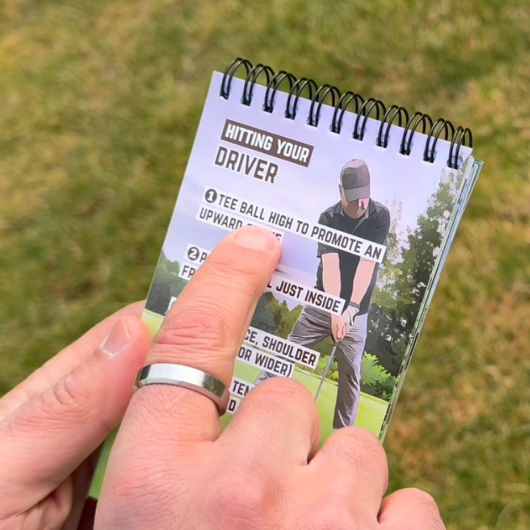 A person holds a spiral-bound golf instruction booklet, pointing to a tip on hitting a driver.