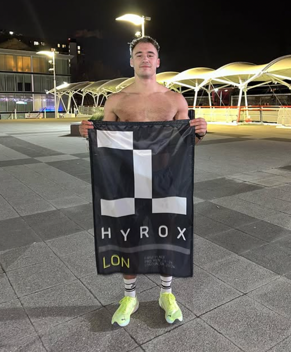Shirtless person holding a 'HYROX LON' banner at night outdoors.
