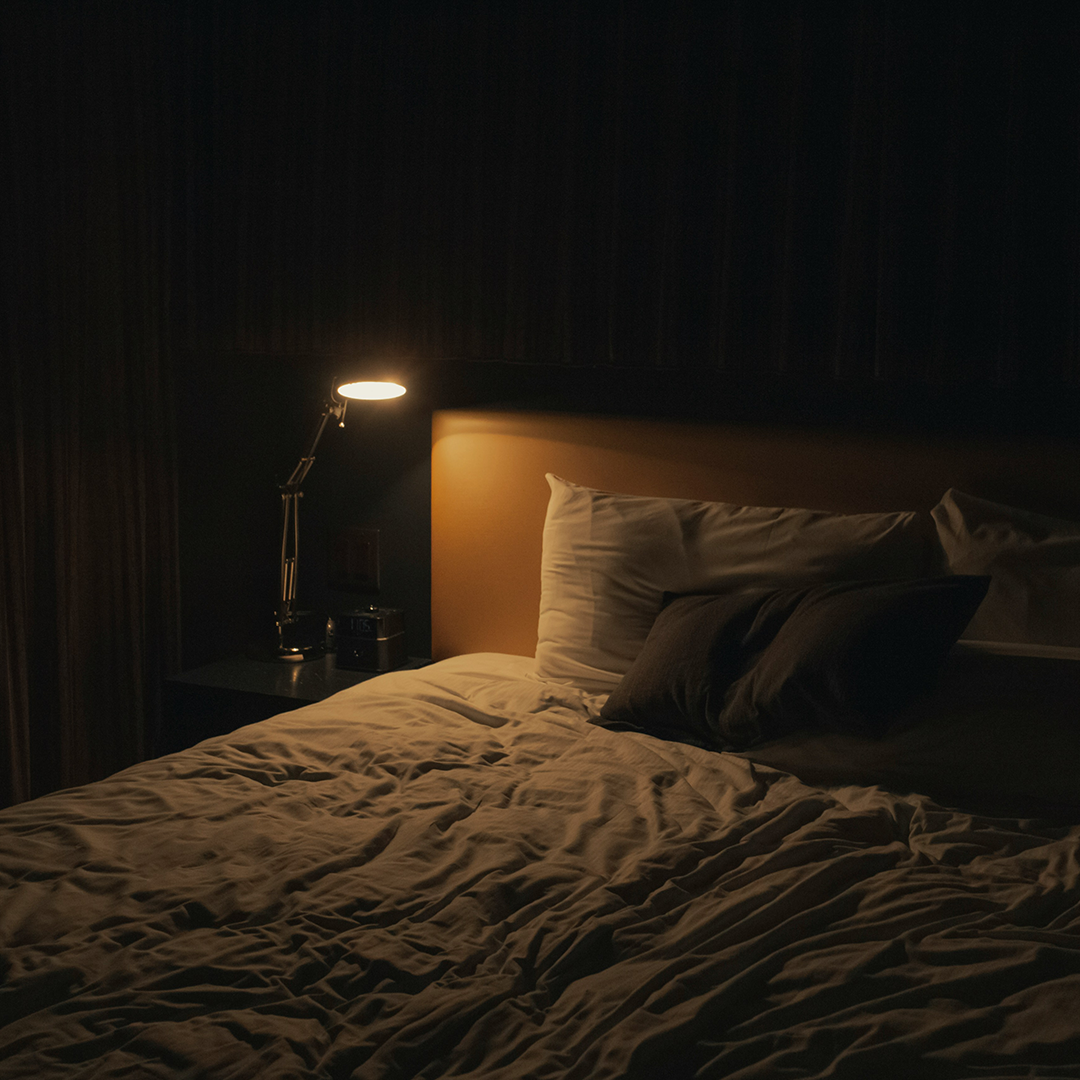 Dimly lit bedroom with a lamp, crumpled sheets, and pillows.