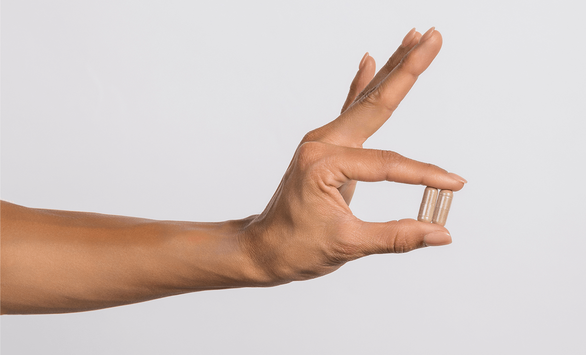 A hand holding two tan capsules between the thumb and forefinger in an 'okay' gesture.