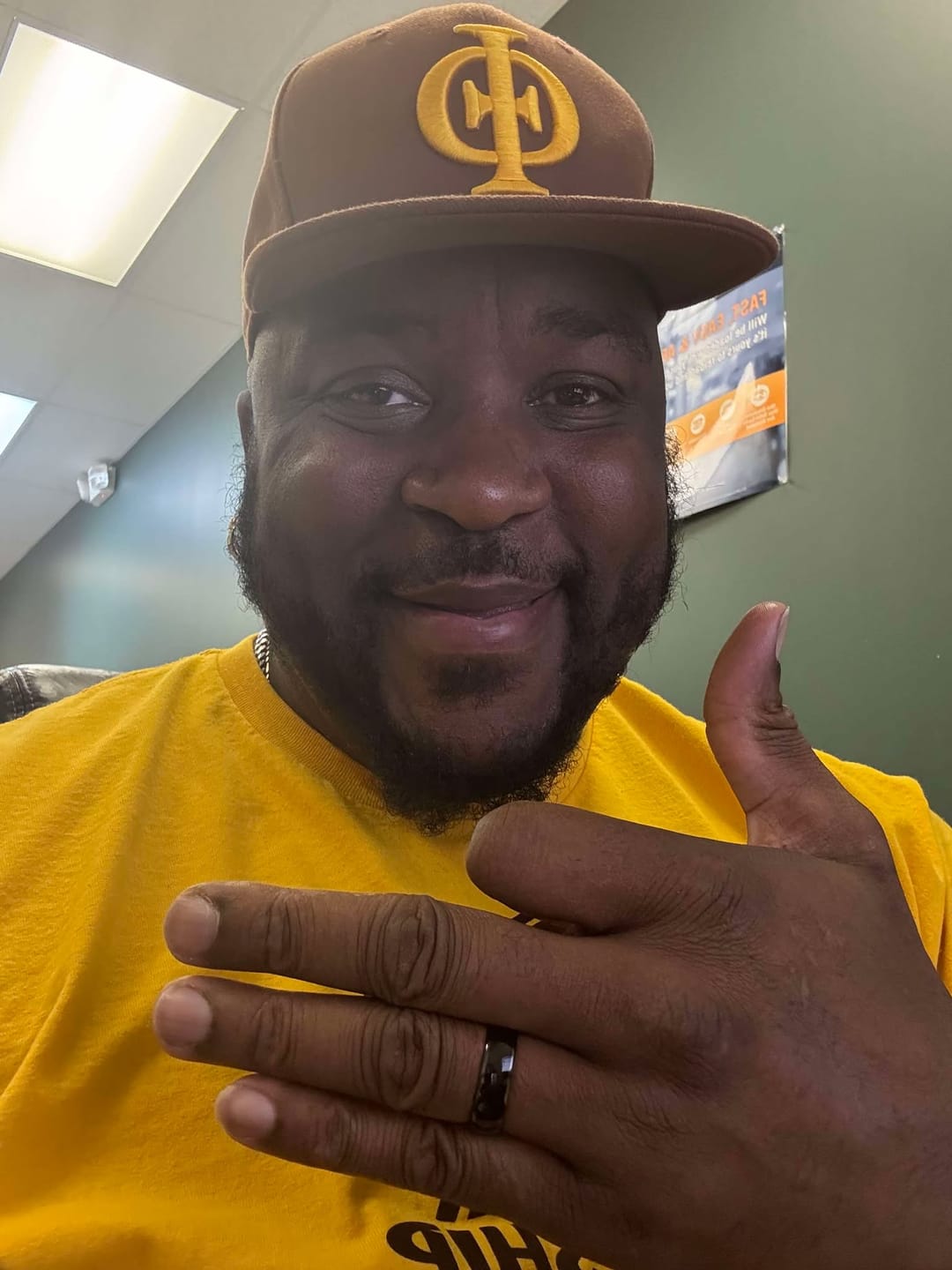 Man smiling and gesturing with hand, wearing a yellow shirt and brown cap indoors.
