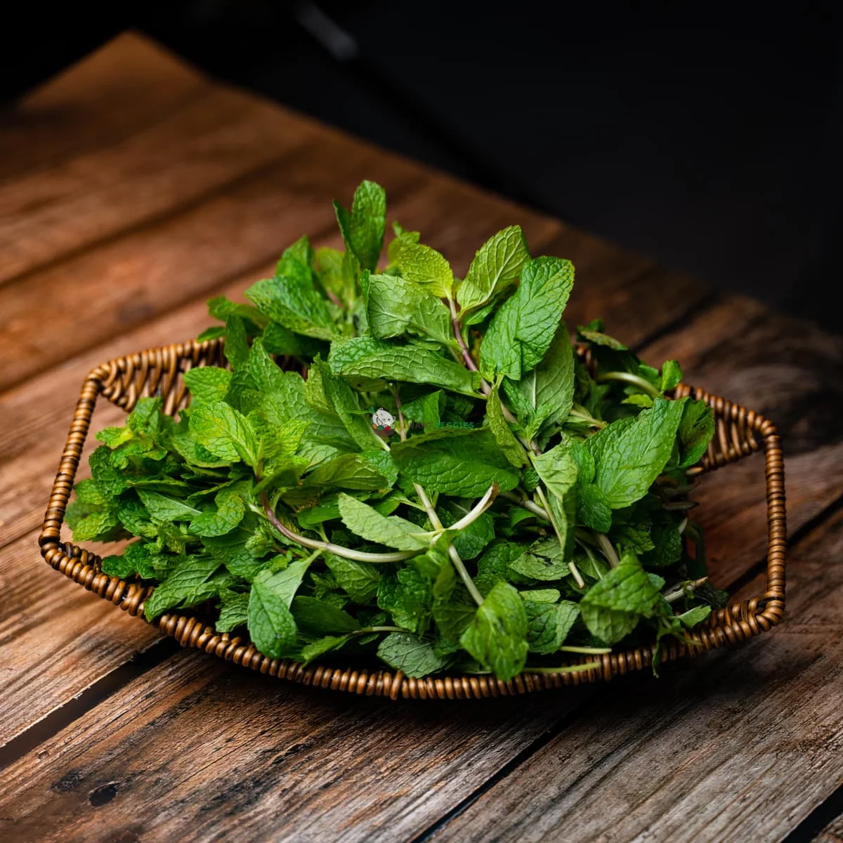 A shallow, woven basket filled with fresh green mint sprigs, sitting on a dark wooden surface.