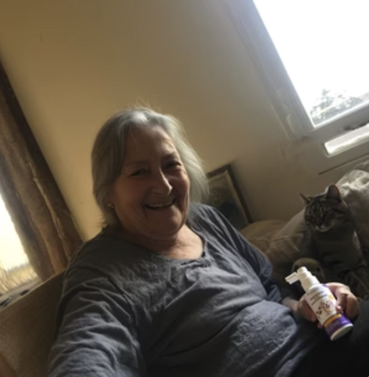 A smiling older woman sits on a couch next to a cat, holding a small spray bottle.