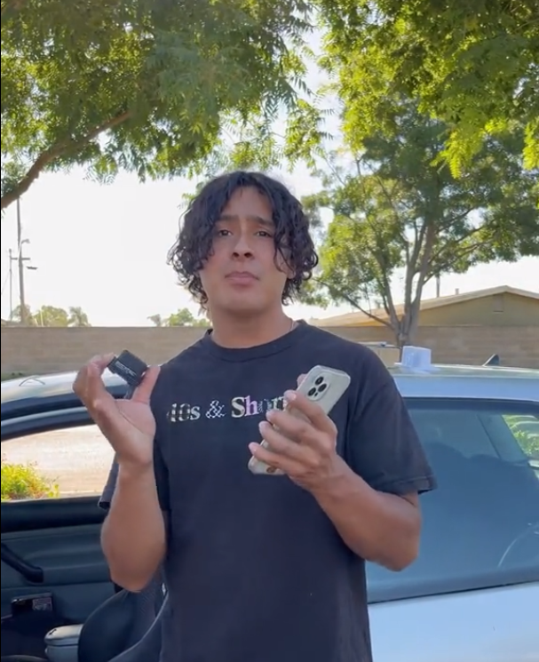 Person holding a phone and a small object near a car, with trees in the background.