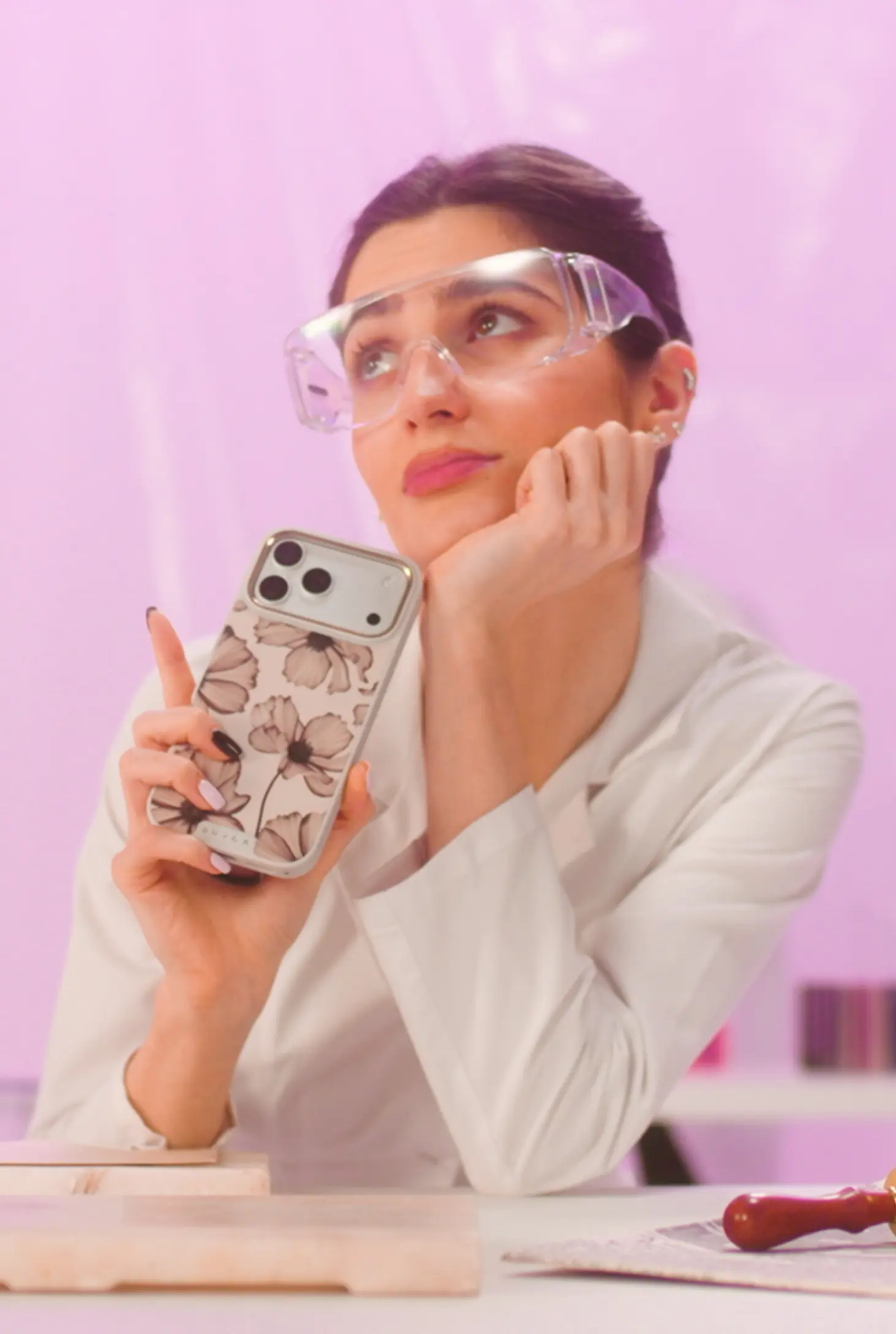 A woman in safety goggles and a white coat holds a floral phone case, looking up thoughtfully.