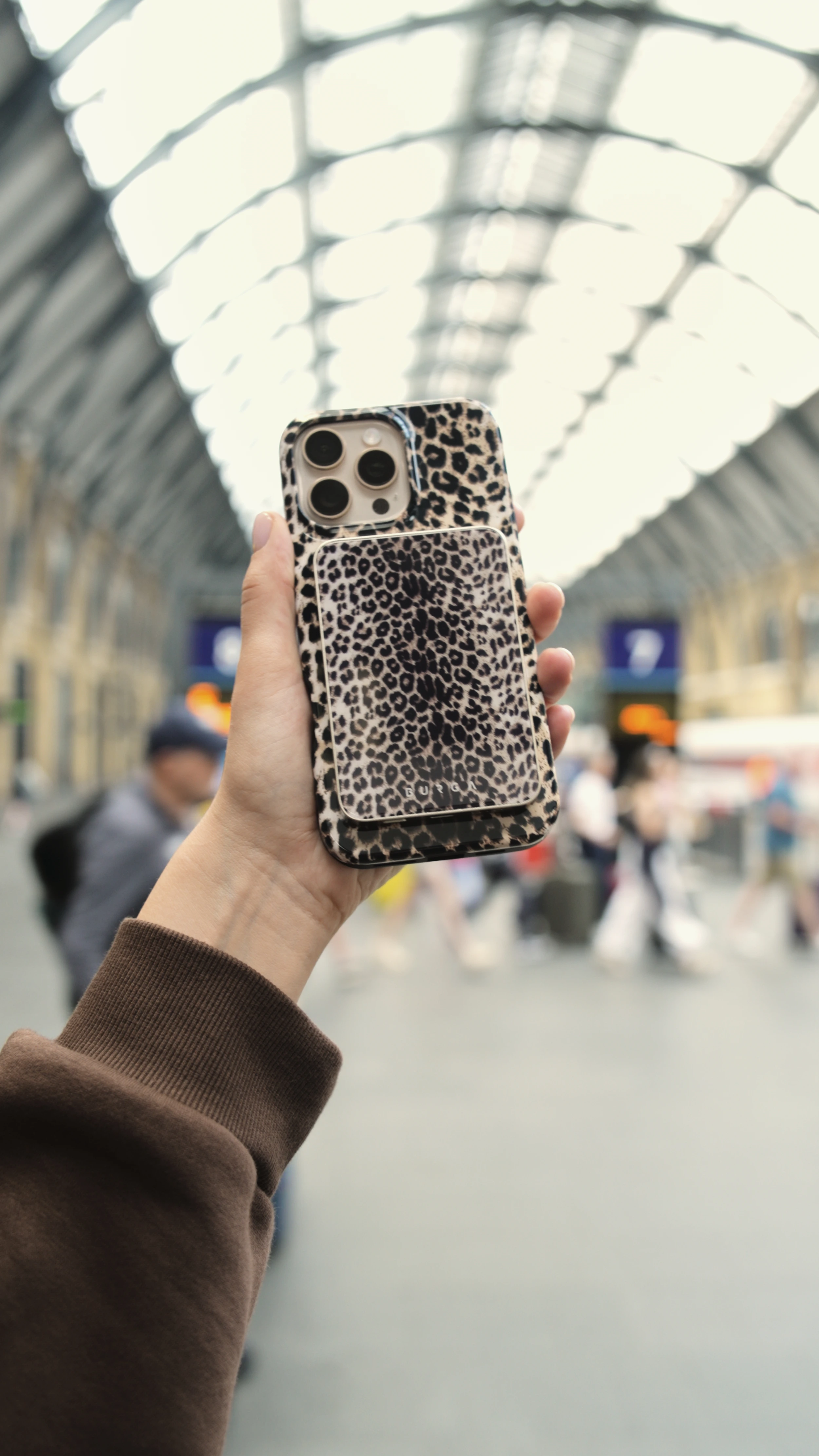 Hand holding a phone with a leopard print case in a busy station.