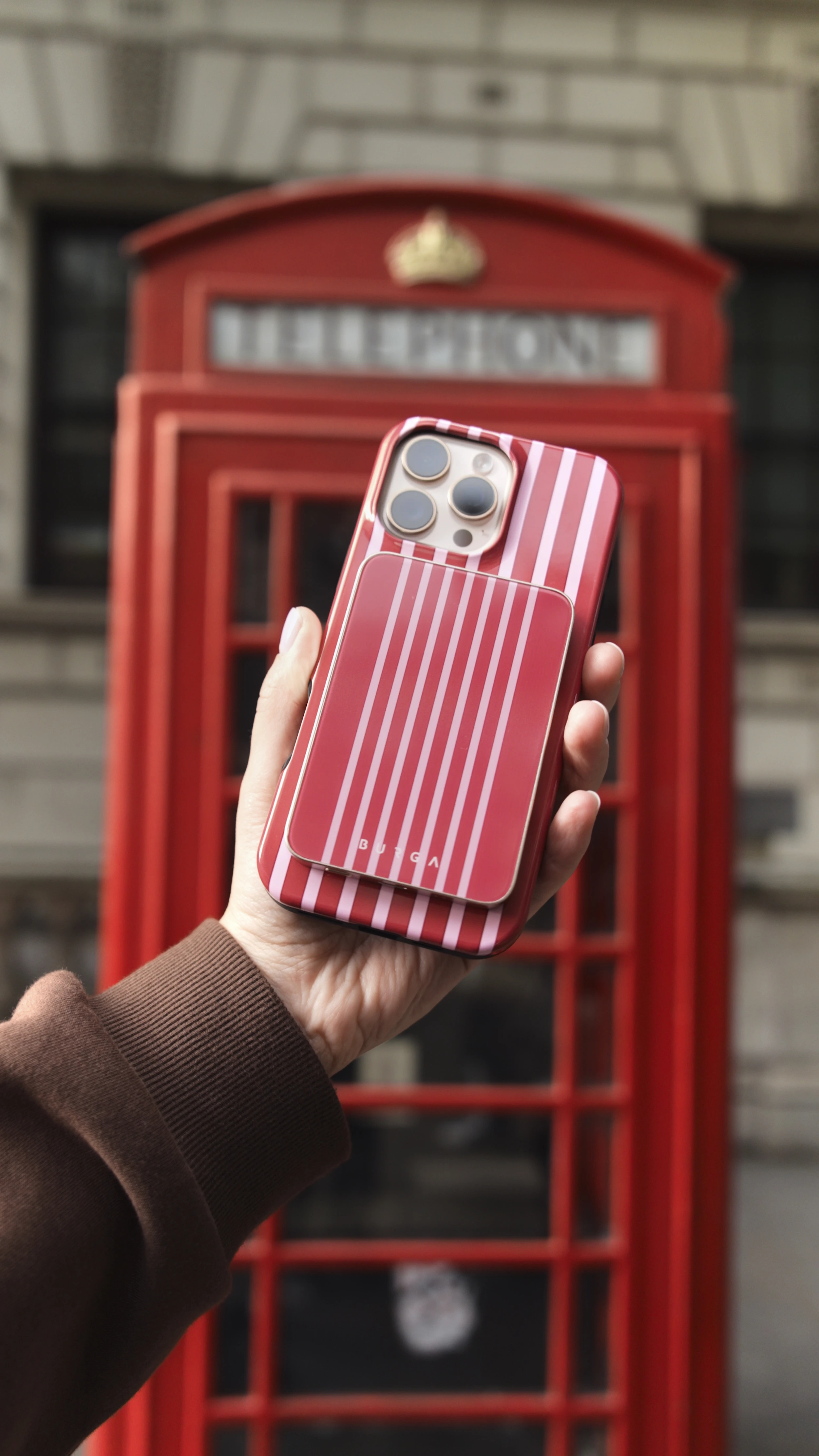 Hand holding a striped phone case in front of a red telephone booth.