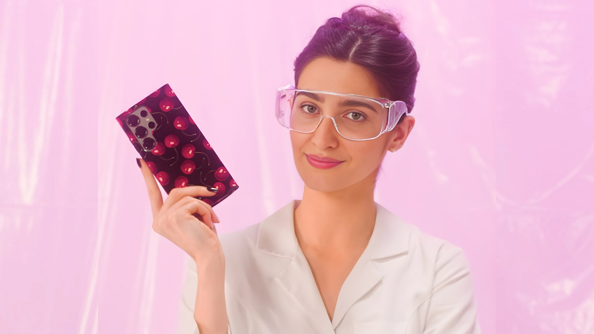 A woman in a lab coat and safety goggles holds up a smartphone with a cherry-patterned case.