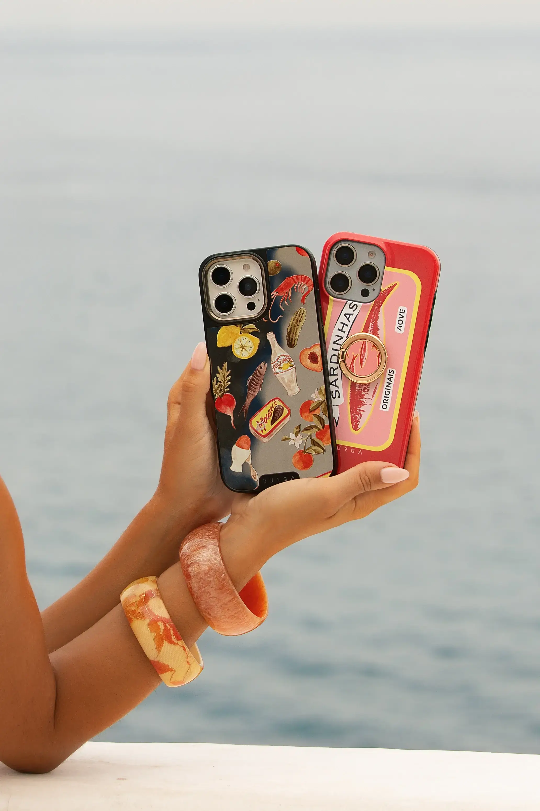 Two hands holding phones with colorful, food-themed cases by the sea.