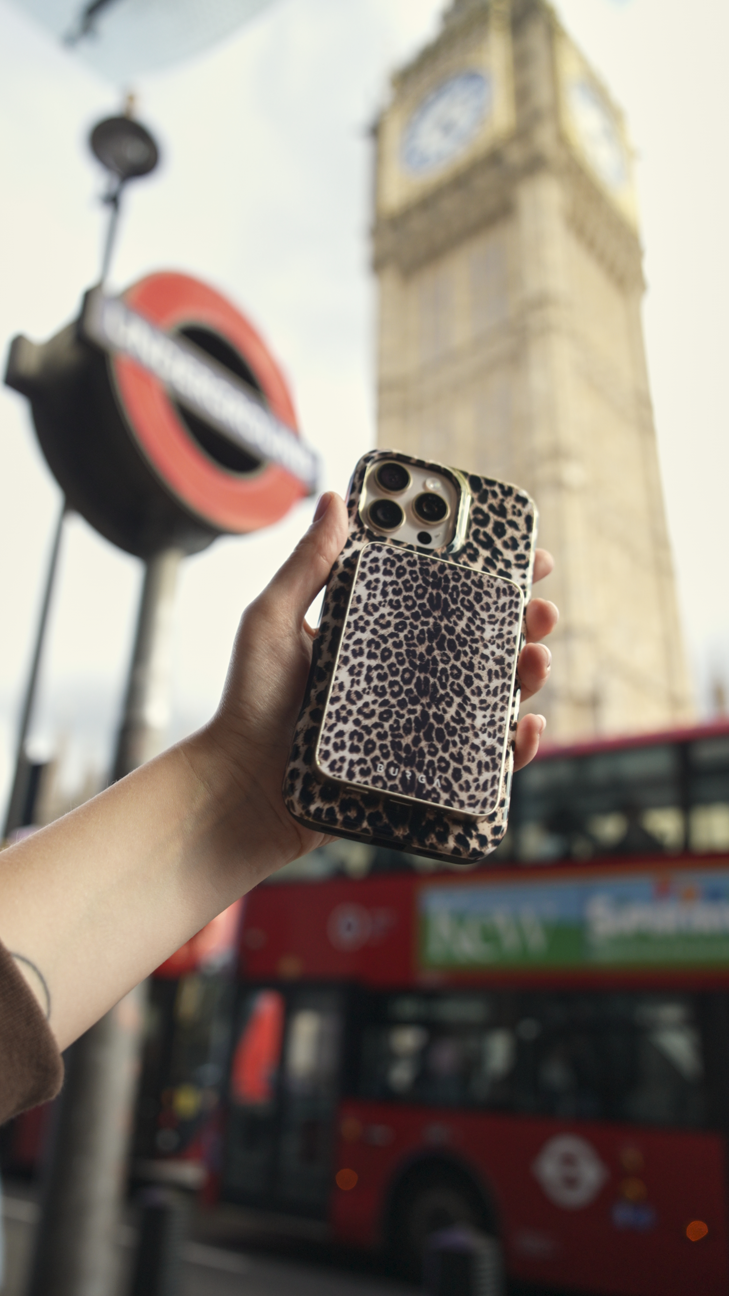 Hand holding leopard print phone case, Big Ben and red bus in background.