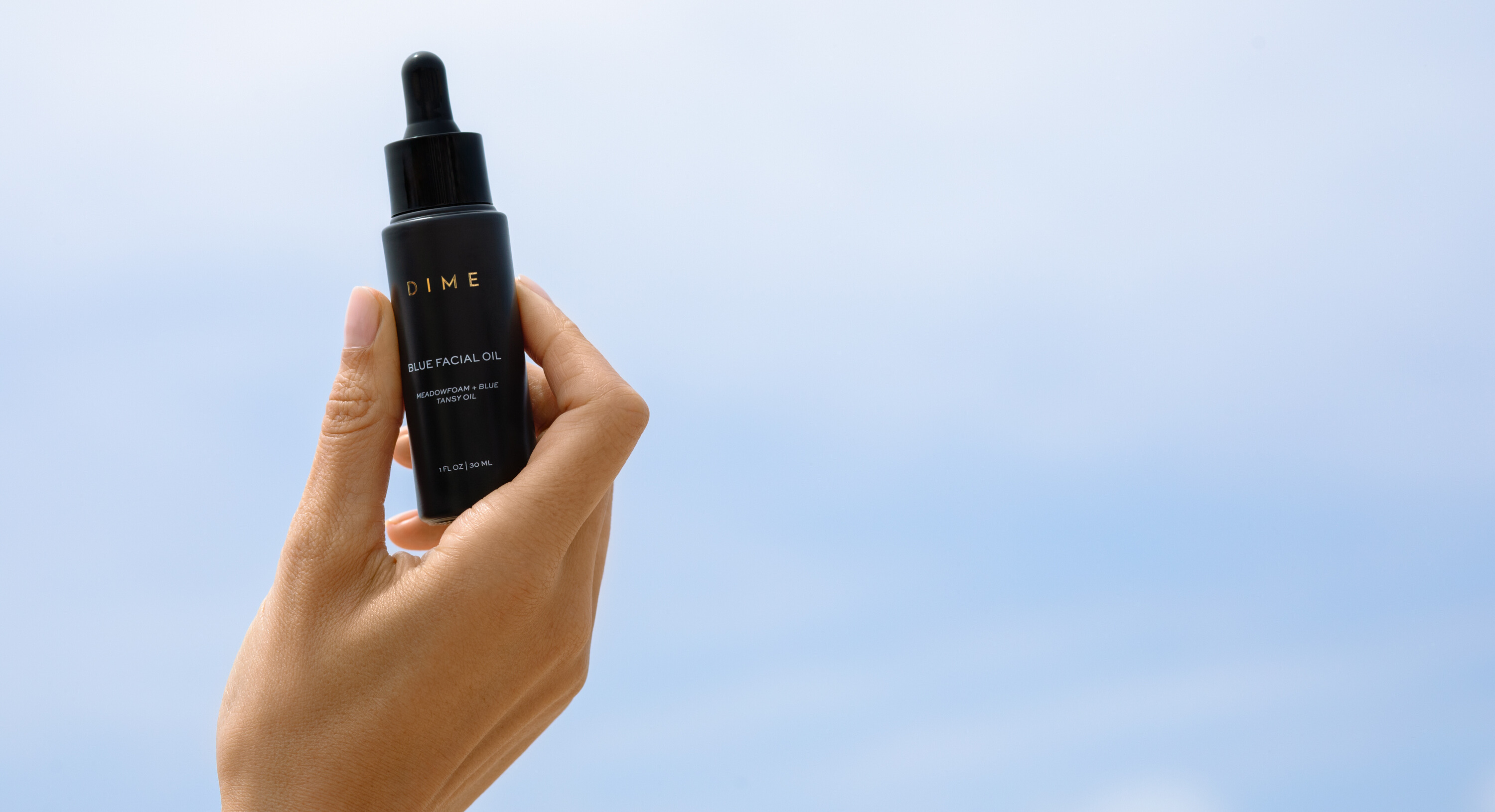 Hand holding a bottle of facial oil against a clear sky.