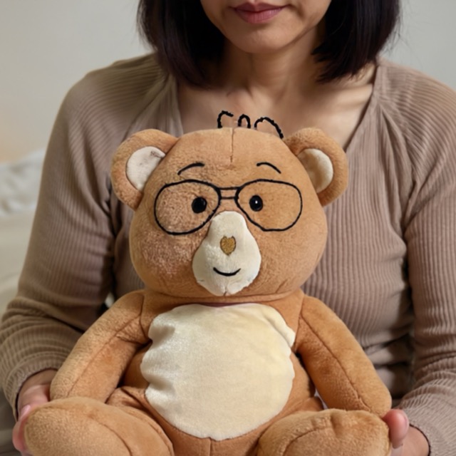 Woman holding a teddy bear with embroidered glasses and a smile.