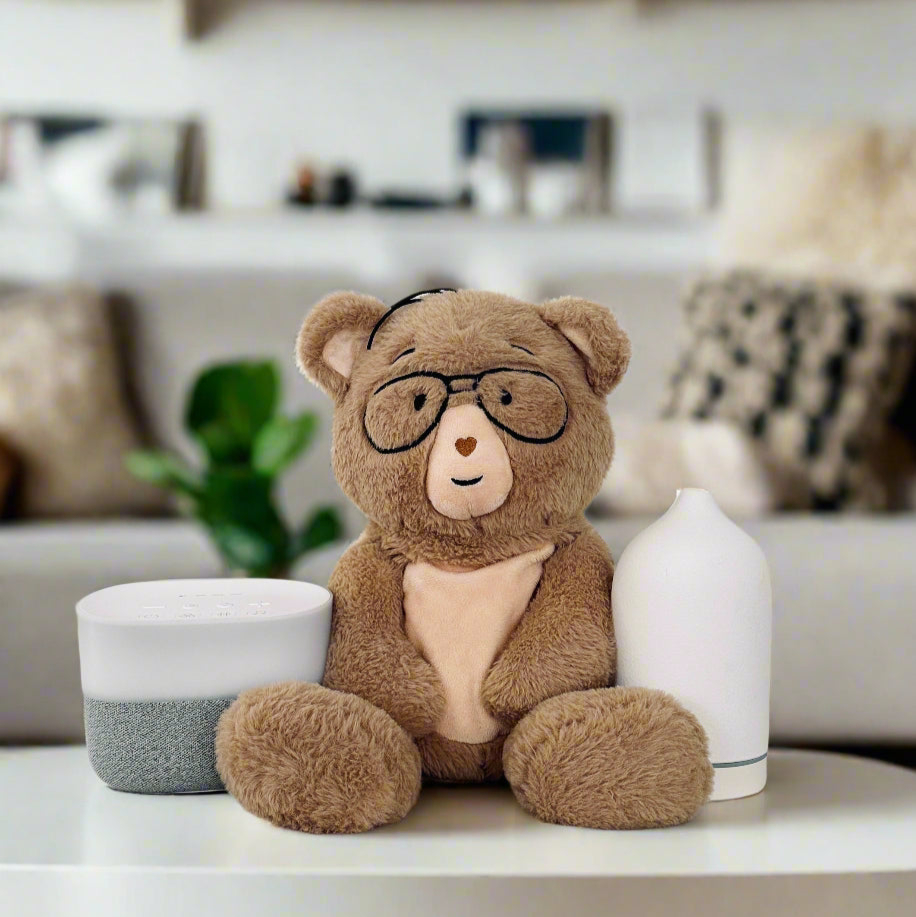 A teddy bear with glasses sits between a speaker and a diffuser on a table.