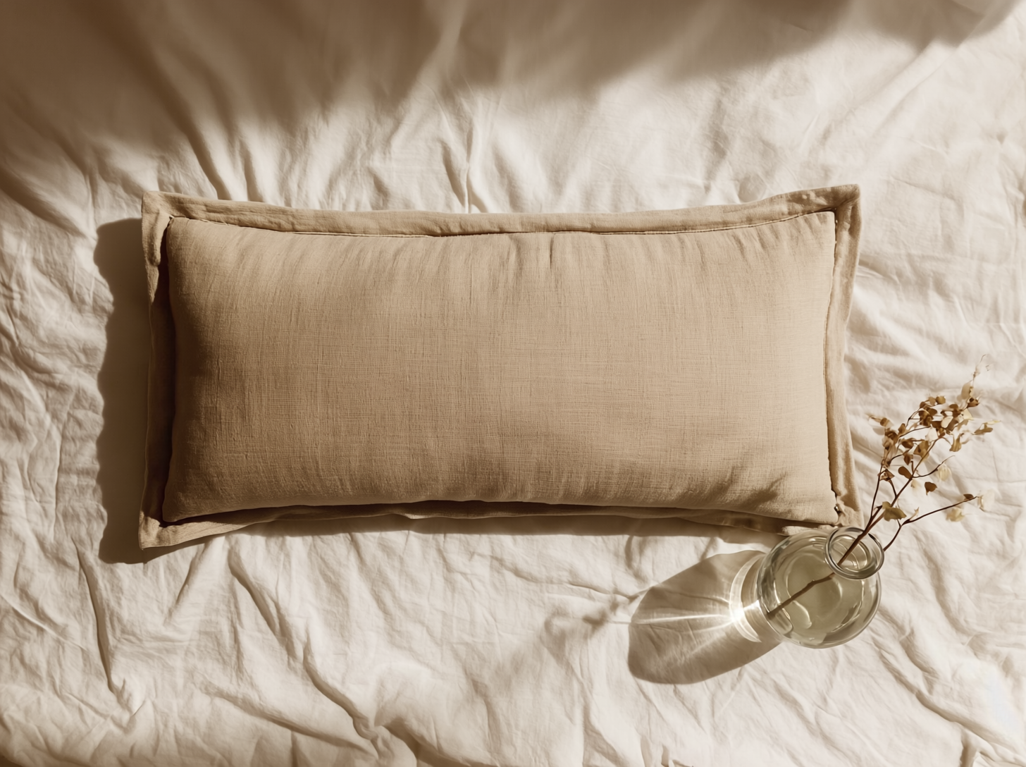 Beige pillow on white bedding with a vase of dried flowers.