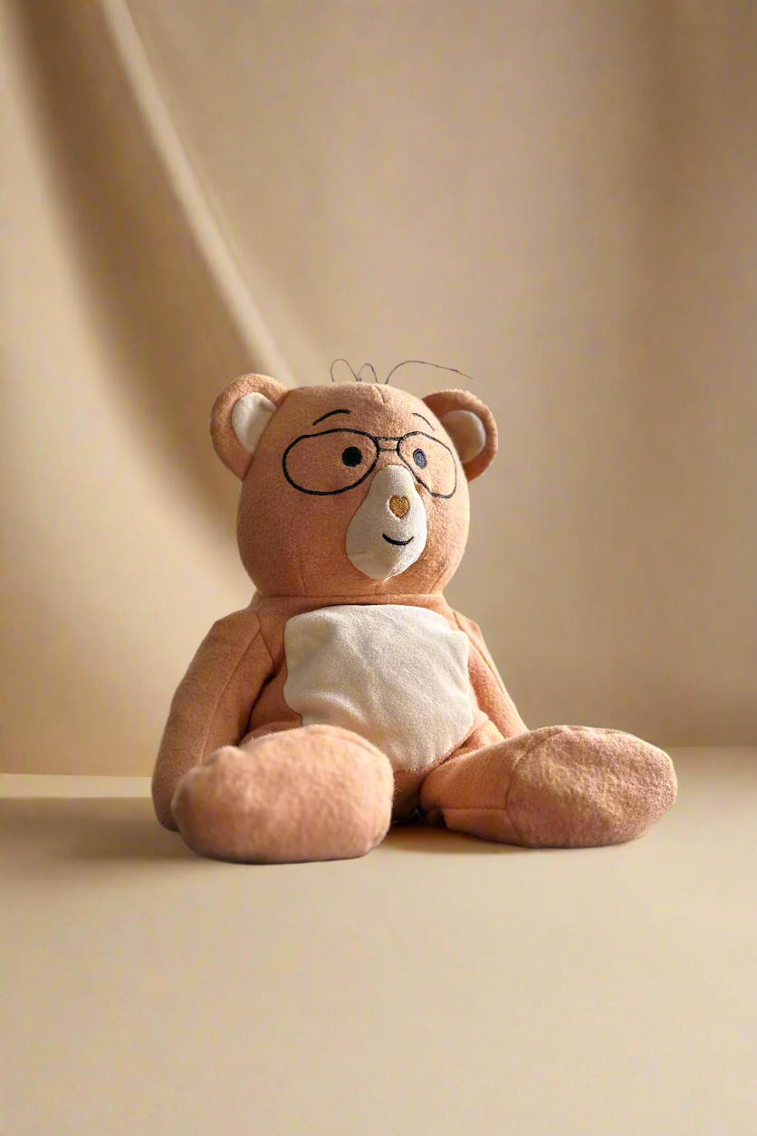 A plush teddy bear with glasses sitting on a beige surface.