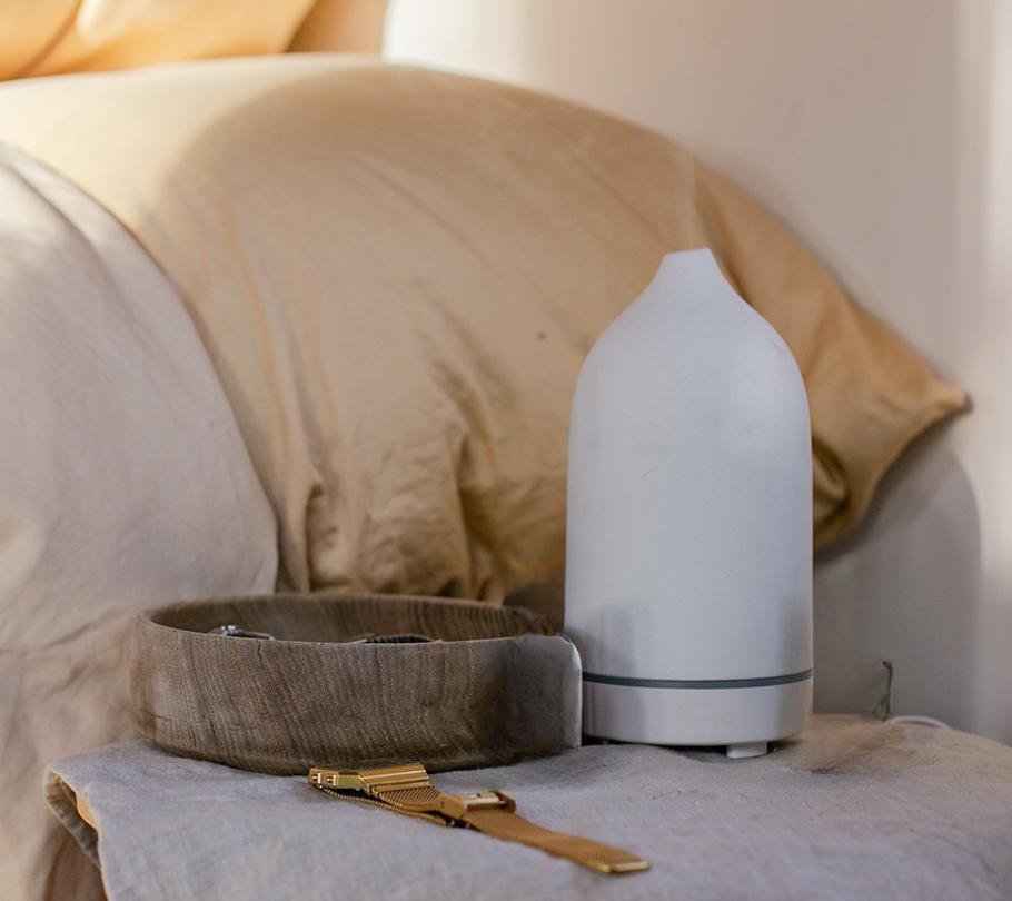 White diffuser and watch on a bedside table with beige bedding.