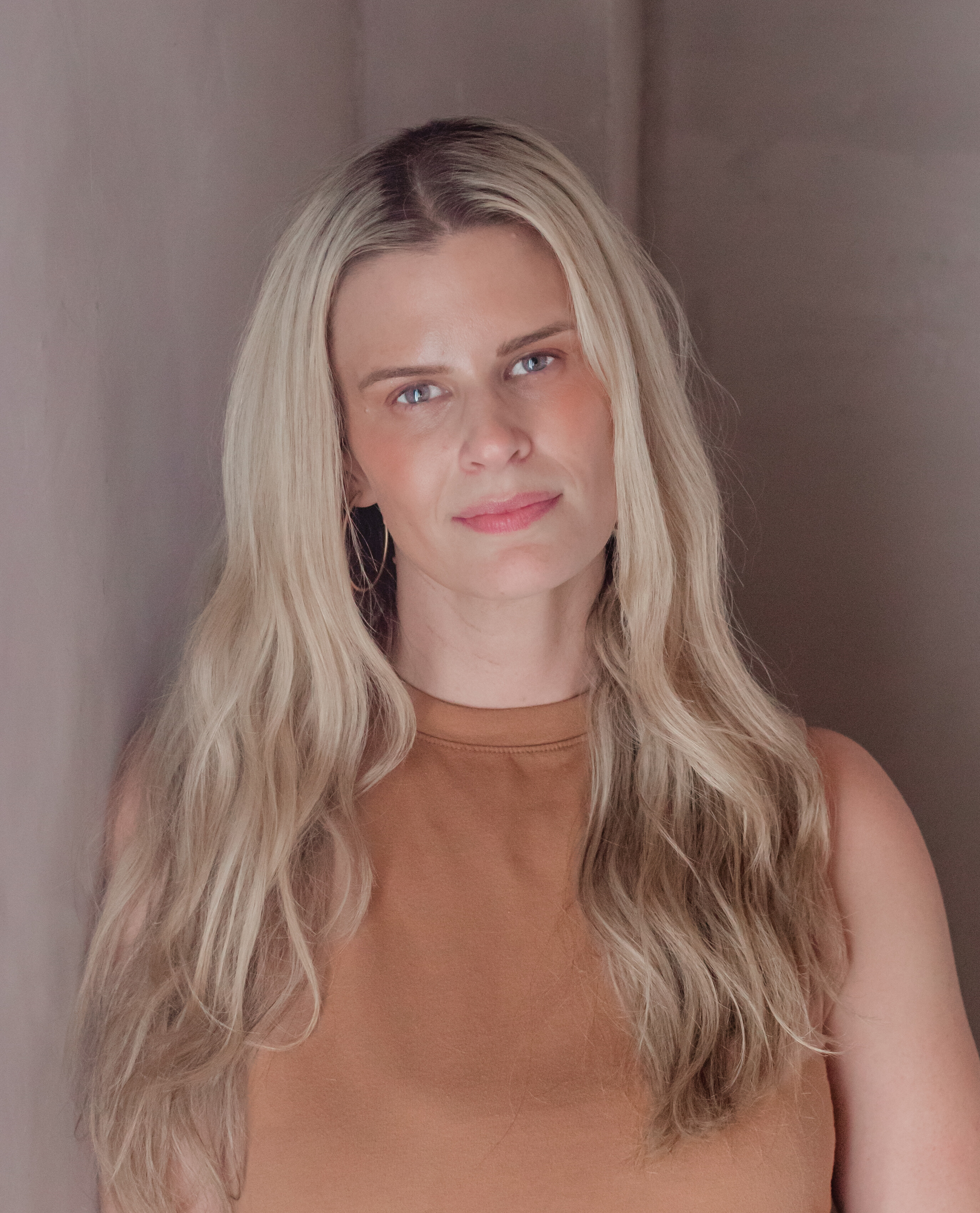 Person with long blonde hair wearing a brown top, standing against a plain wall.
