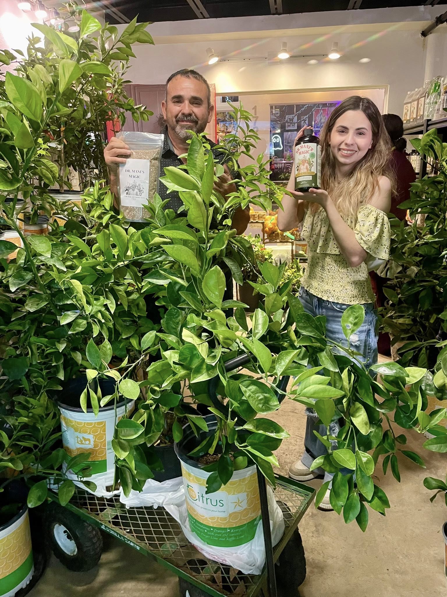 A man and a woman stand in a nursery holding plant care products, surrounded by potted citrus trees.