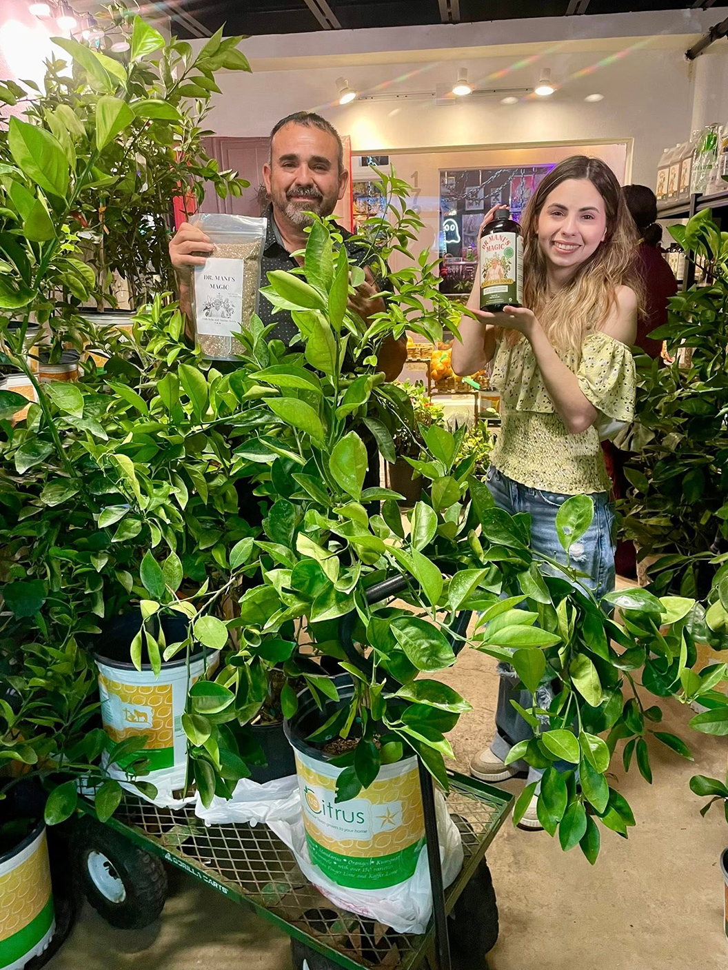 A man and a woman in a plant nursery hold up products while surrounded by potted citrus trees.