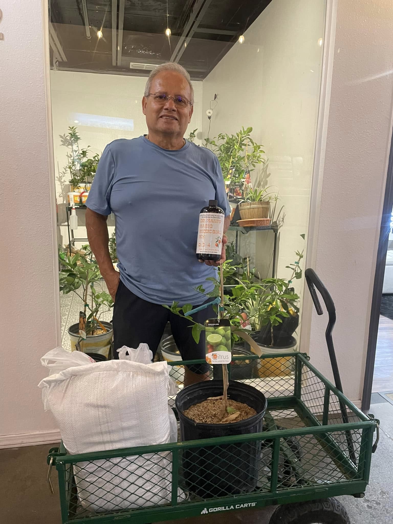 A man stands with a green cart holding a potted citrus plant, a large white bag, and a fertilizer bottle.