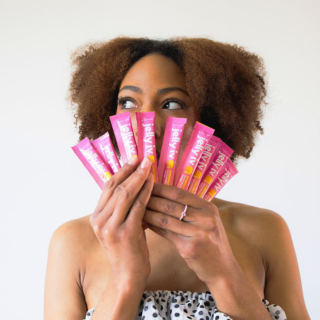 A woman with curly hair holds a fan of pink Jelly IV packets in front of her face.