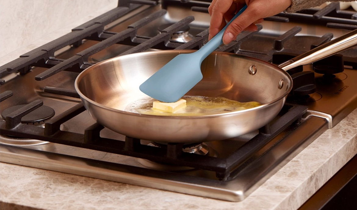 A person uses a blue spatula to melt a pat of butter in a stainless steel skillet.