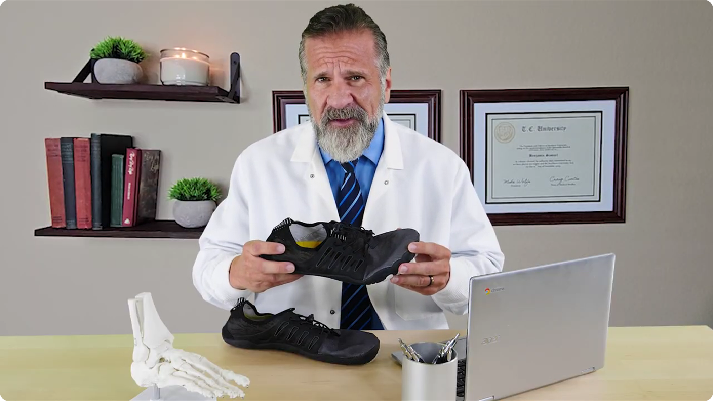 Man in white coat holding black shoes with a laptop on a desk.