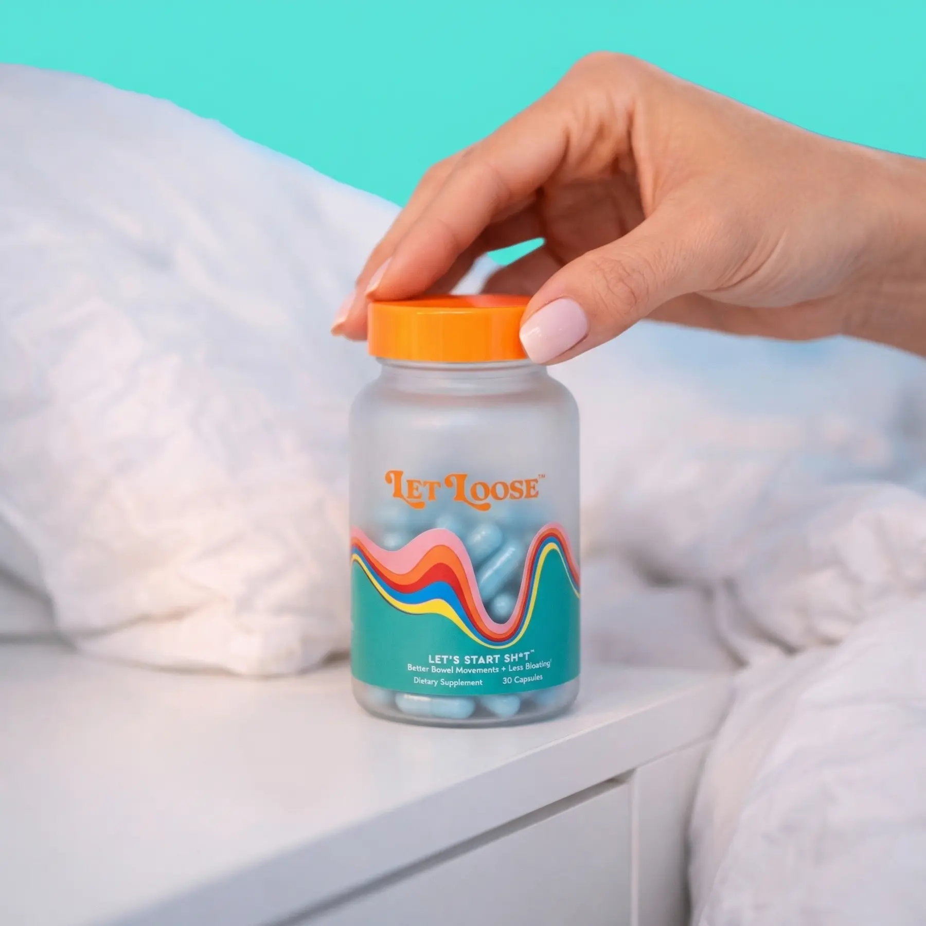 A hand reaches for a bottle of Let Loose dietary supplements on a white nightstand next to a bed.