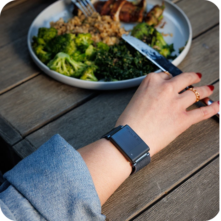A hand with a smartwatch near a plate of broccoli and other food on a wooden table.