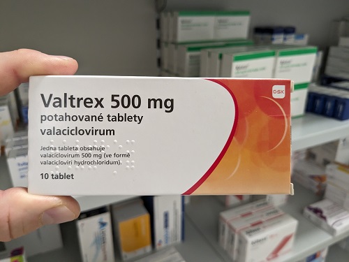 A person's hand holding a box of Valtrex 500 mg medication in front of pharmacy shelves.