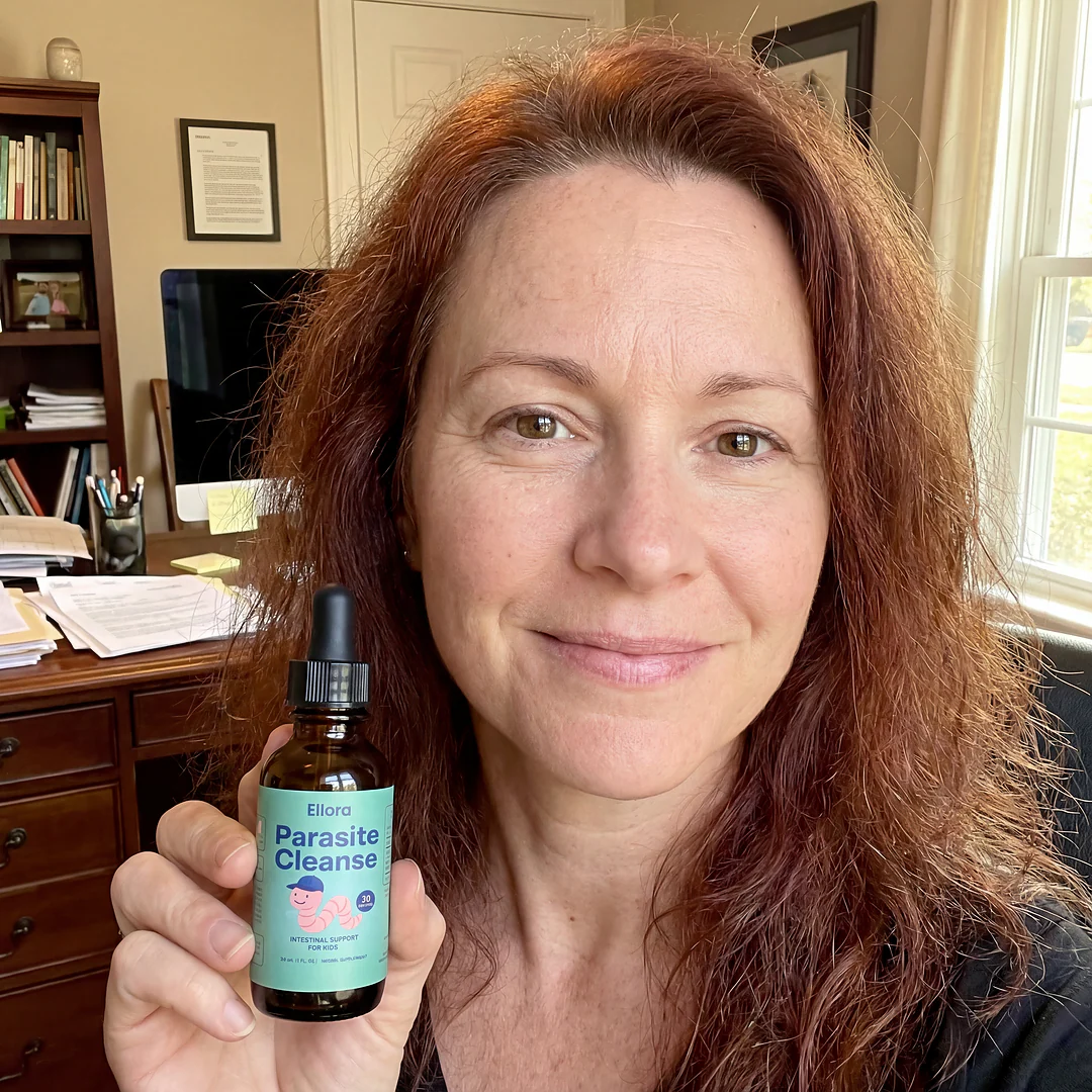 A woman with red hair holds up a small bottle of Ellora Parasite Cleanse in an office.