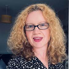 Person with curly blonde hair and glasses smiling indoors.