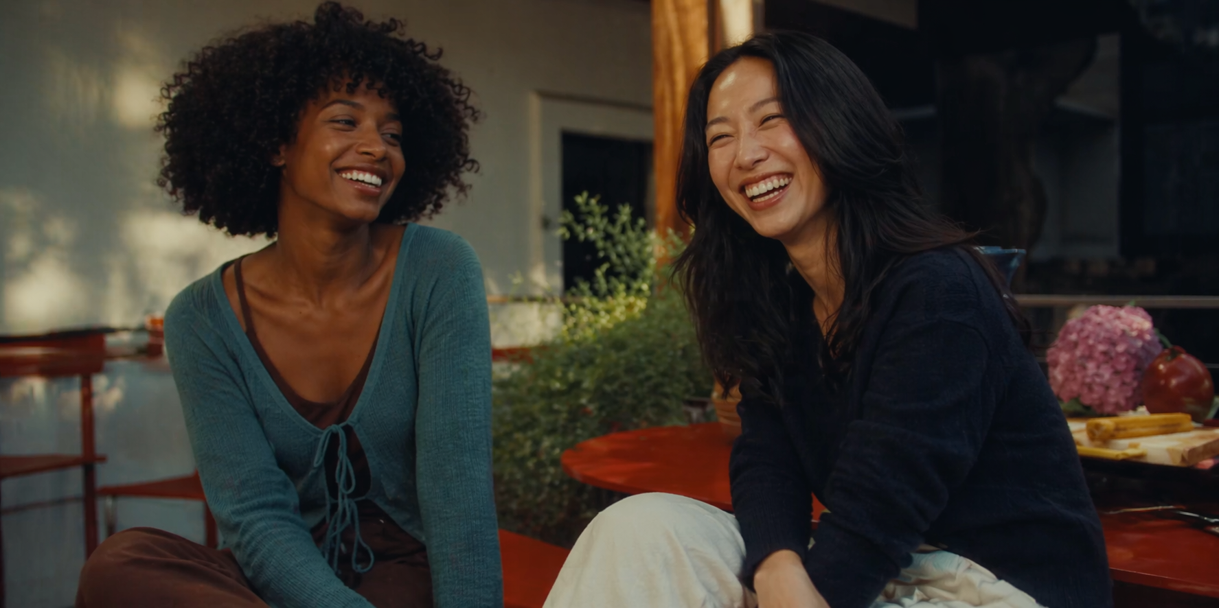 Two women, one Black and one East Asian, are sitting together and laughing outdoors.