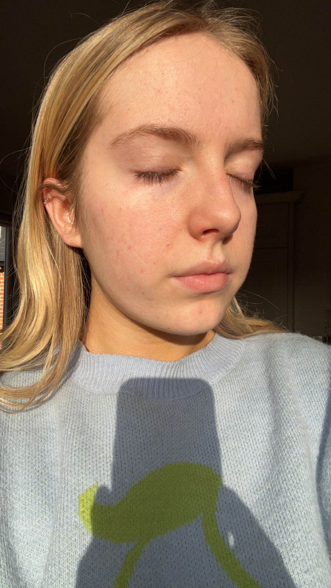 A young person with blonde hair stands in the sun with their eyes closed, wearing a light blue sweater.
