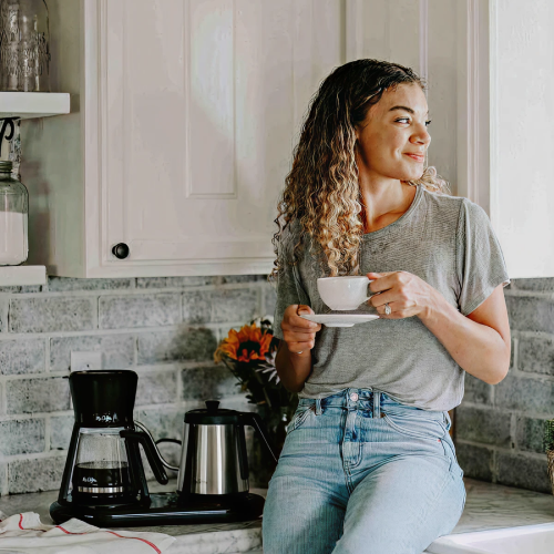 Woman sitting on kitchen counter holding a coffee cup, with a coffee maker nearby.