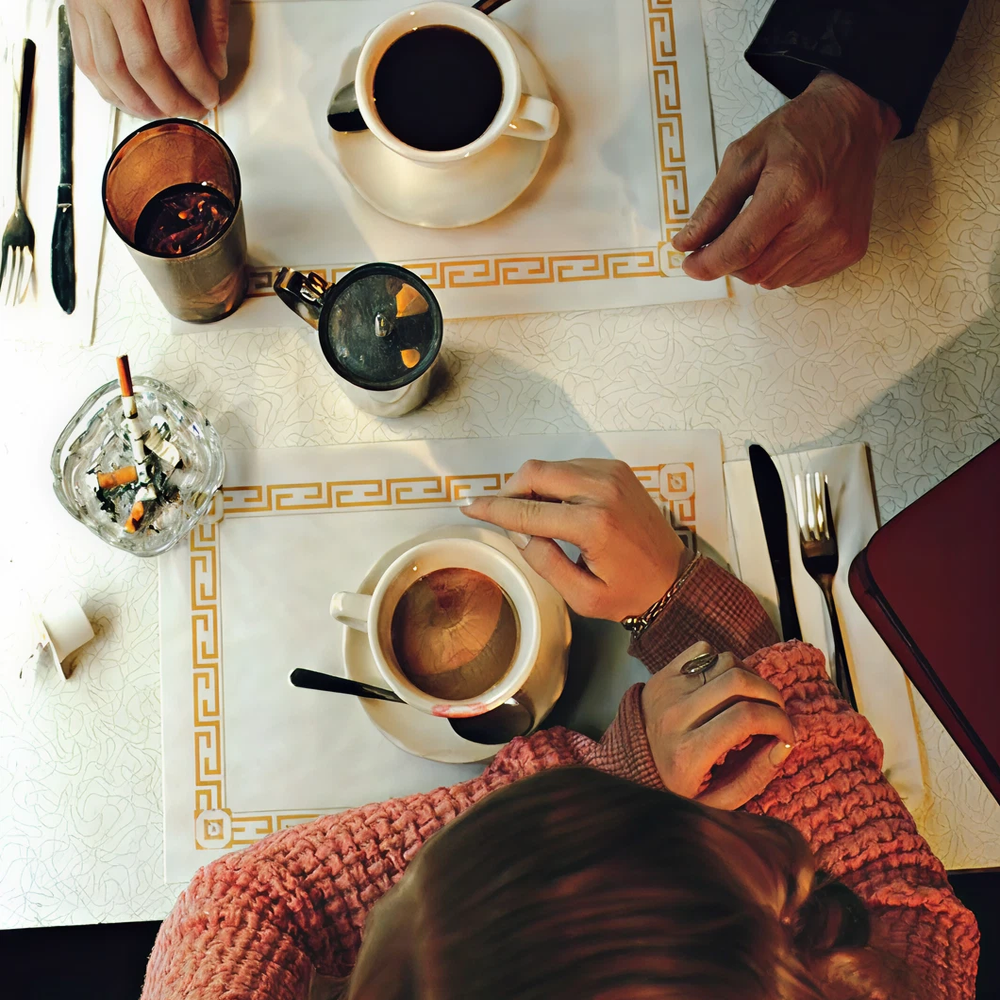 Two people at a table with coffee, drinks, and an ashtray.