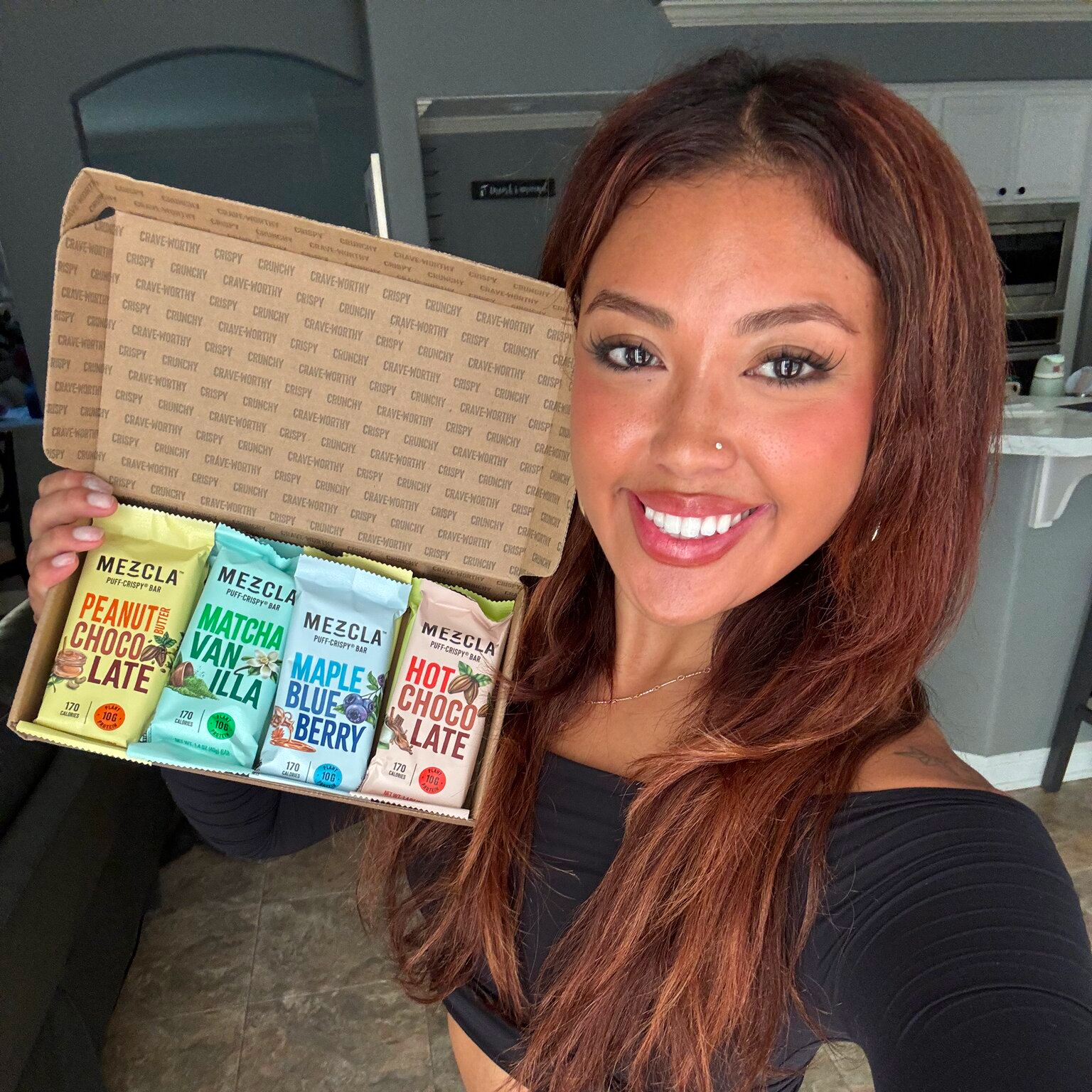 Person holding a box of assorted Mezcla protein bars, smiling indoors.