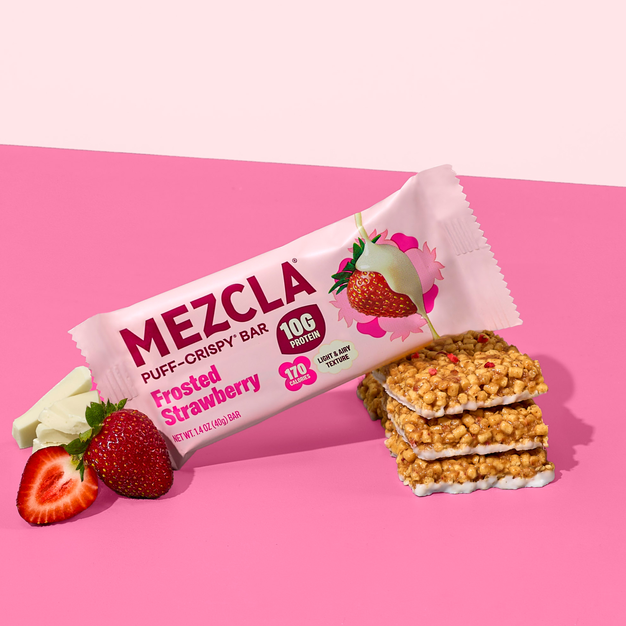 A Mezcla Frosted Strawberry protein bar with unwrapped bars, fresh strawberries, and white chocolate on a pink background.