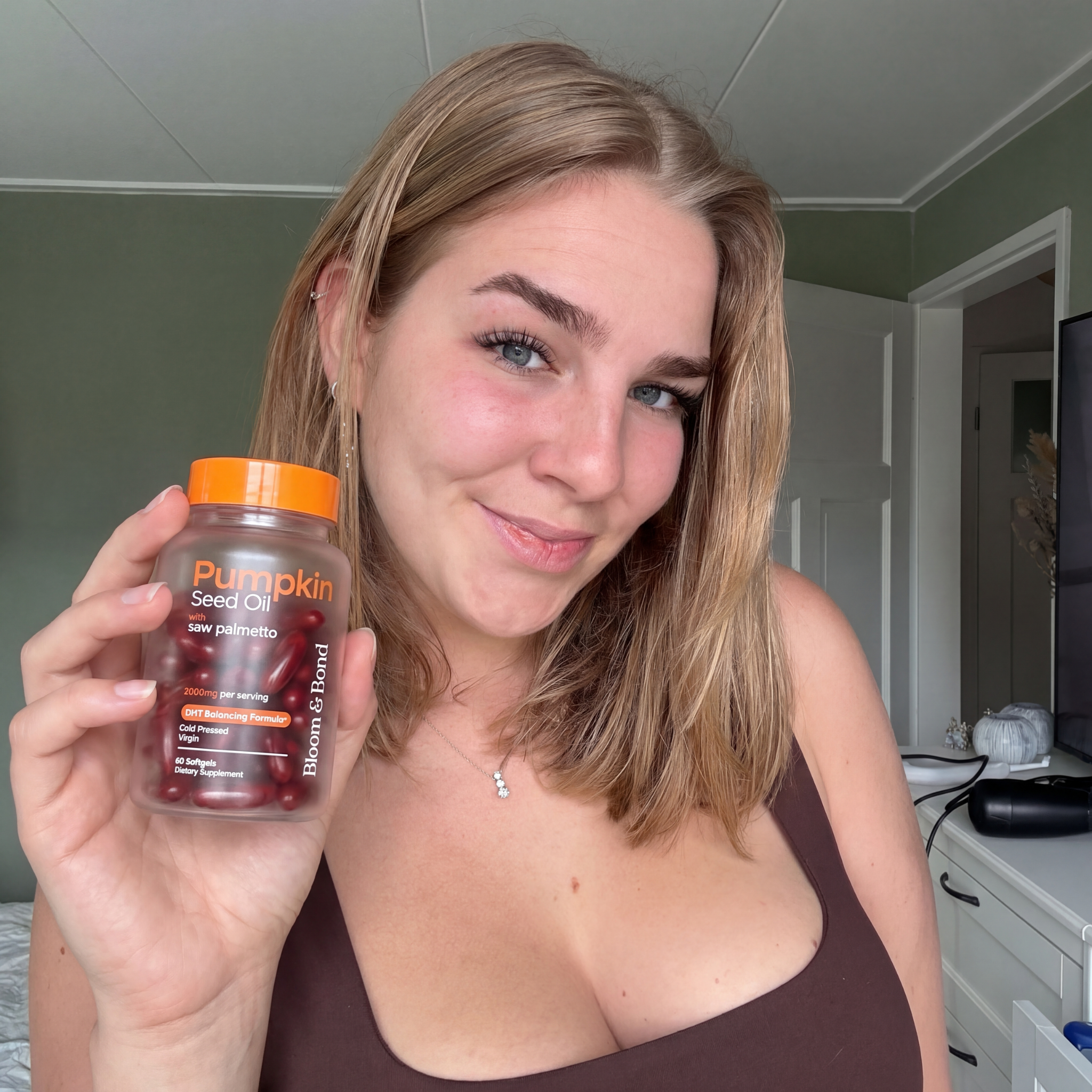 A smiling woman holds up a bottle of Bloom & Bond Pumpkin Seed Oil softgels in a bedroom.