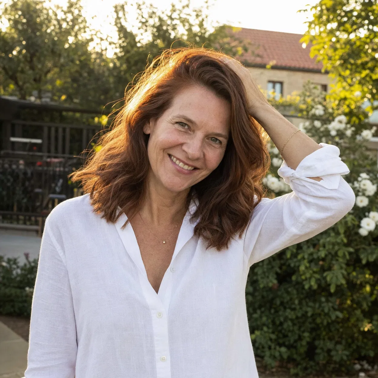 A smiling woman with auburn hair and a white shirt stands outside with her hand in her hair.