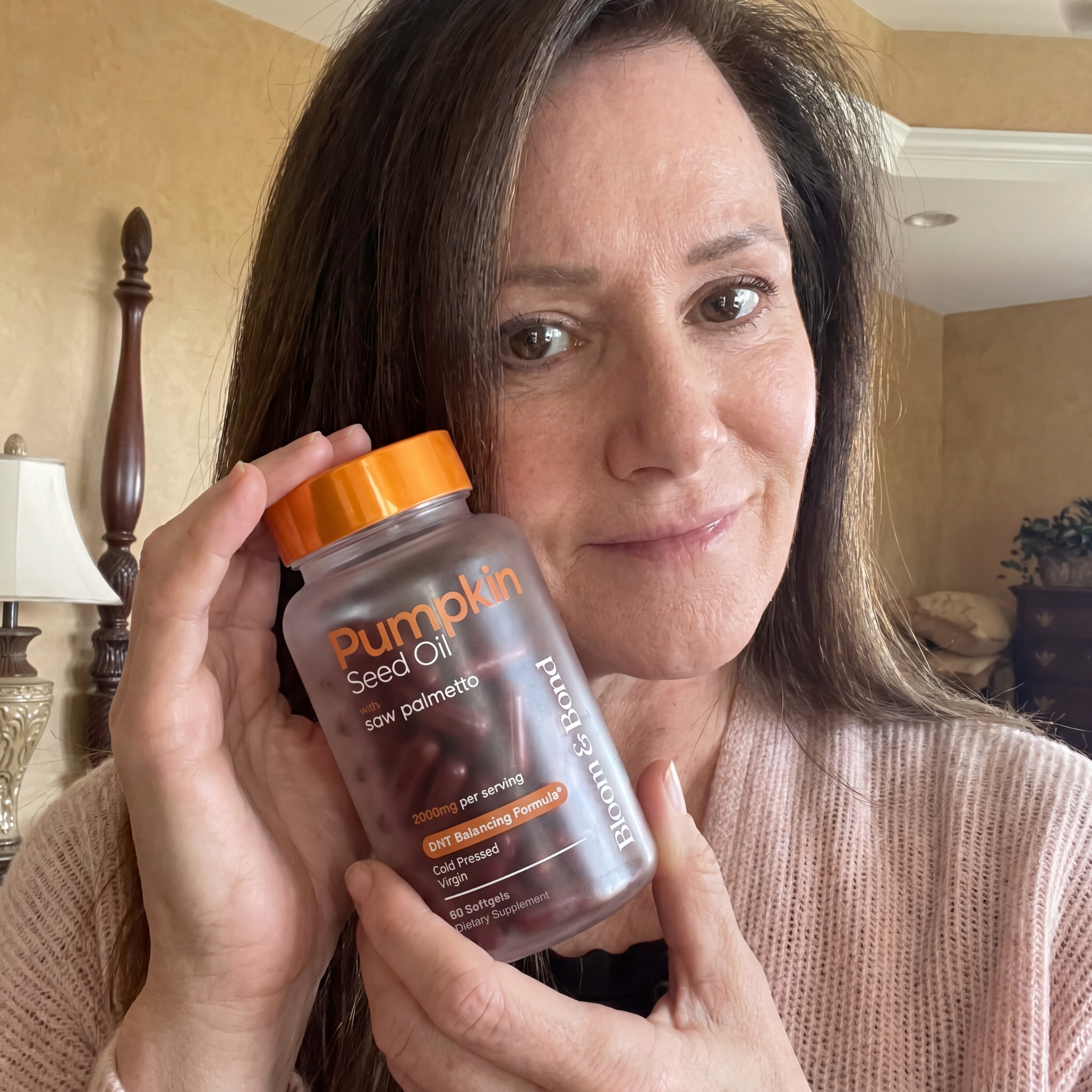 A woman with brown hair holds a bottle of Pumpkin Seed Oil supplements next to her face and smiles.